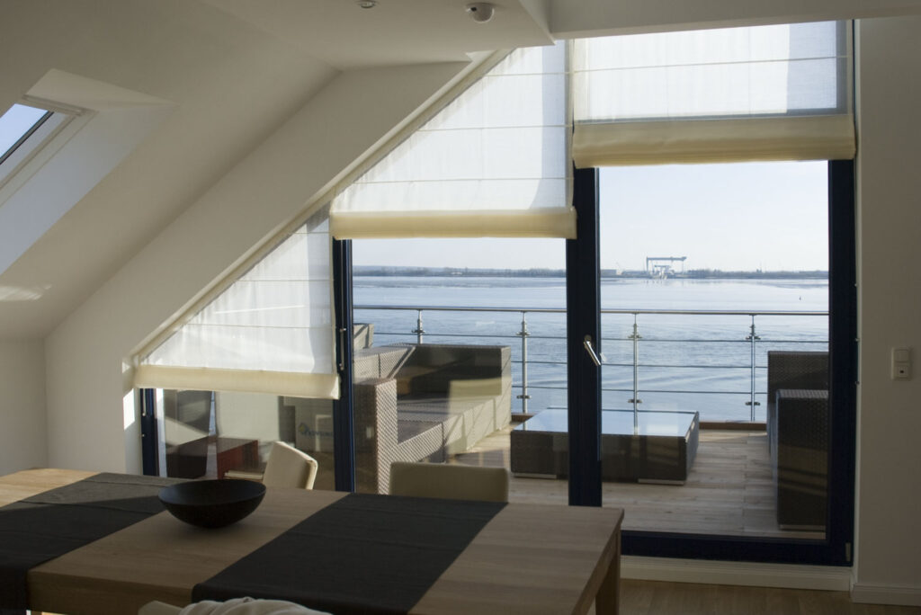 Maßgefertigte Biesenfaltrollos für schräge und gerade Fenster, elegant kombiniert mit modernem Wohndesign und Blick auf das Wasser.