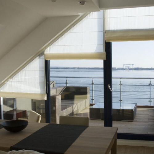 Maßgefertigte Biesenfaltrollos für schräge und gerade Fenster, elegant kombiniert mit modernem Wohndesign und Blick auf das Wasser.