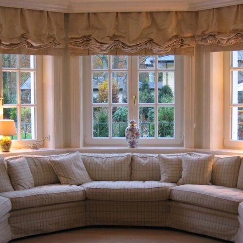 Elegantes Wohnzimmer mit großen Sprossenfenstern und mehreren üppigen Mailänder Raffrollos nebeneinander in heller Farbgebung.