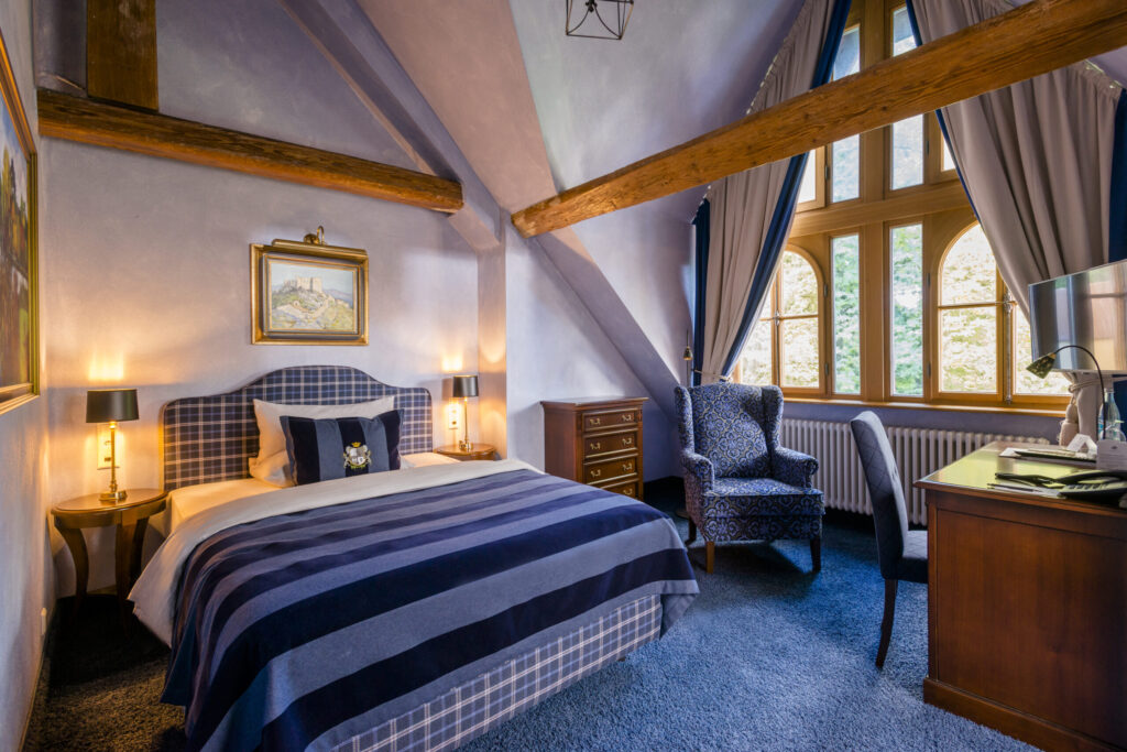 Elegantes Schlosshotel-Zimmer mit gemütlichem Ambiente, rustikalen Holzbalken und maßgefertigten, abgeschrägten Vorhängen.