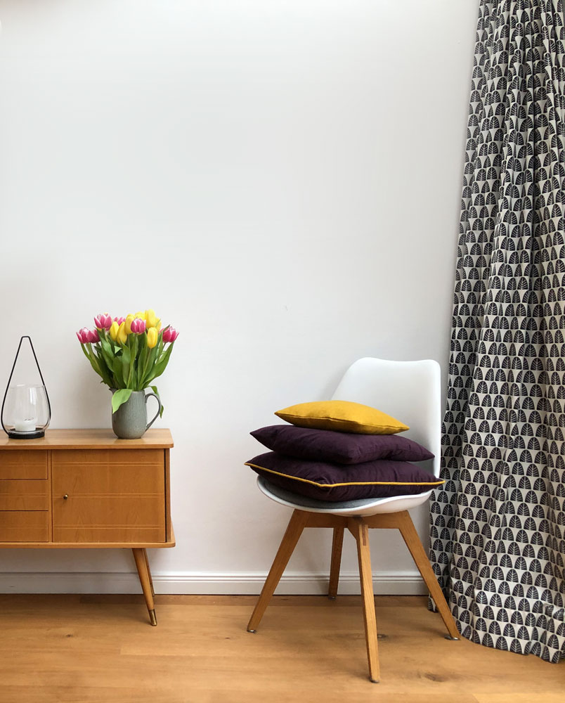 Skandinavisches Interieur mit Retro-Sideboard, gemustertem Vorhang und dekorativen Kissen auf einem Designerstuhl.
