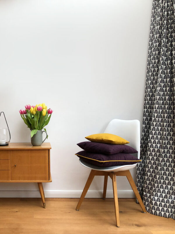 Skandinavisches Interieur mit Retro-Sideboard, gemustertem Vorhang und dekorativen Kissen auf einem Designerstuhl.