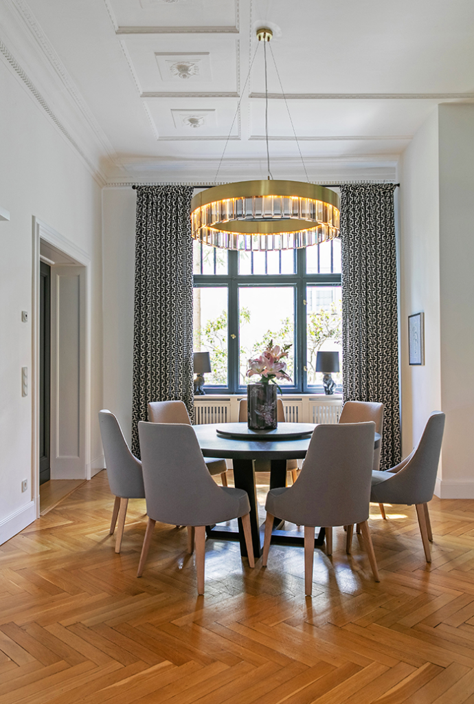 Elegantes Esszimmer mit rundem Tisch, gepolsterten Stühlen, opulentem Kronleuchter und Vorhänge mit Ringen an einer Stilstange.