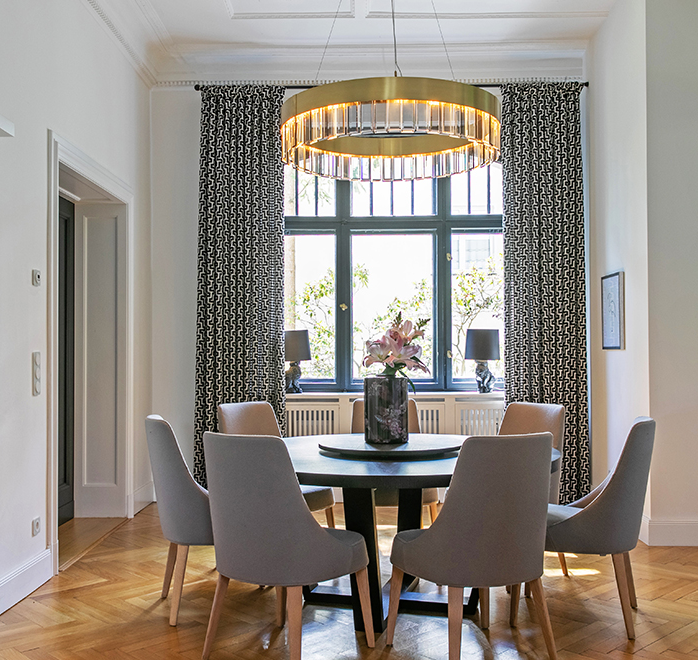 Elegantes Esszimmer mit rundem Tisch, gepolsterten Stühlen, opulentem Kronleuchter und Vorhänge mit Ringen an einer Stilstange.