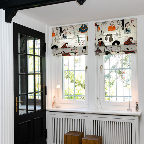 Quadro Faltrollos mit kunstvollem, buntem Muster in einem stilvollen Wohnraum mit weißen Fensterrahmen und Holzelementen.