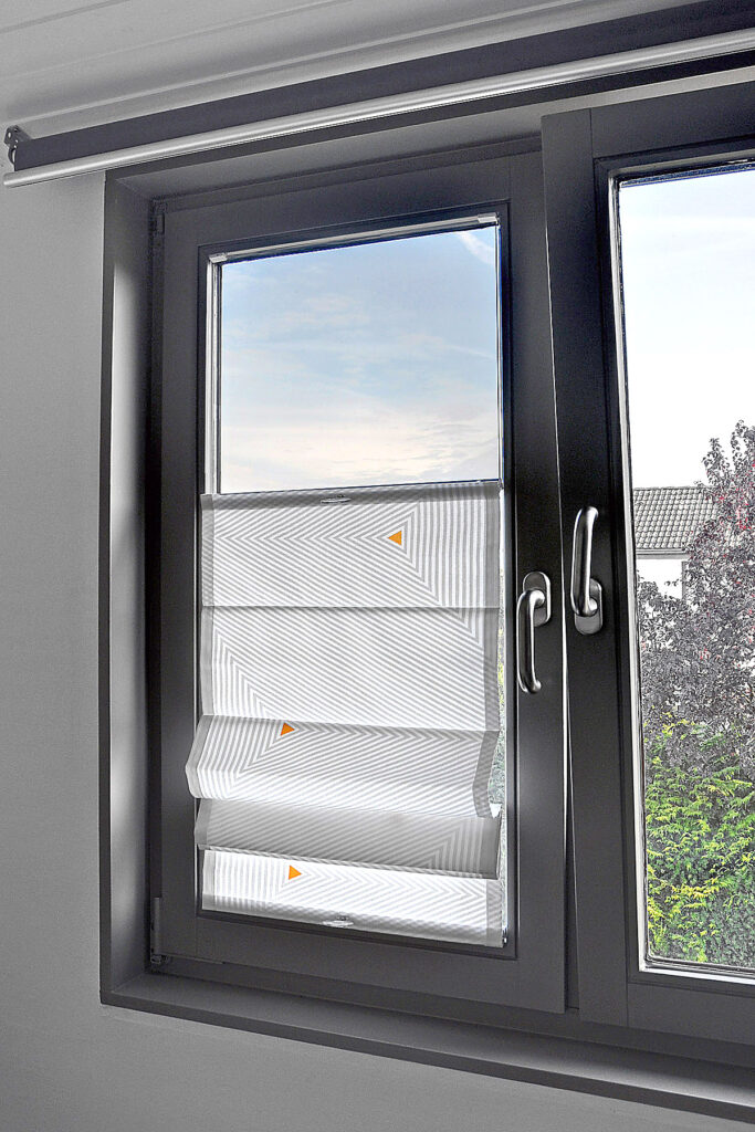 Faltrollo zum Verschieben mit geometrischem Muster in Grau und kleinen farblichen Akzenten, montiert an einem modernen Fenster mit dunklem Rahmen.