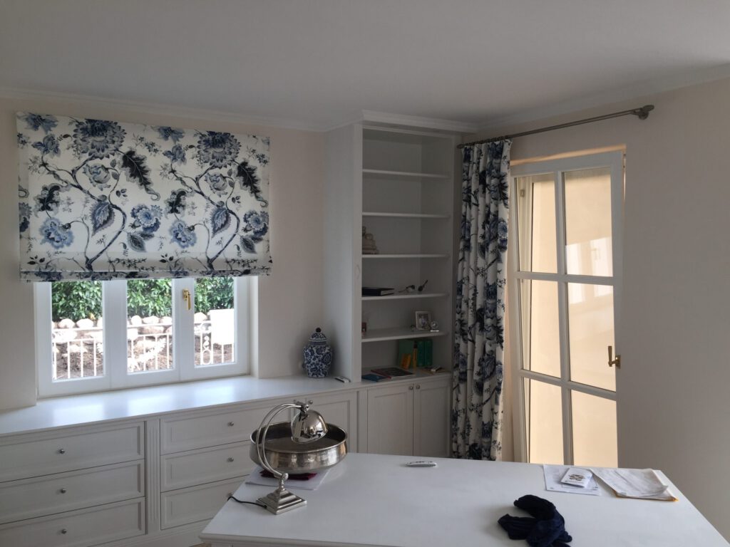 Faltrollo und Vorhang mit blau-weißem Blumenmuster vor weißen Fenstern in einem hellen, klassisch eingerichteten Raum mit Einbauschränken und Bücherregal.